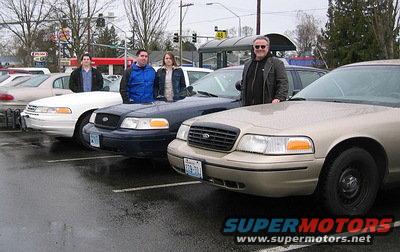 Four Ford Crown Victoria sedans parked in a lot. Three men and one woman stand by the cars