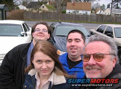 Four people pose for a selfie with three cars visible behind them