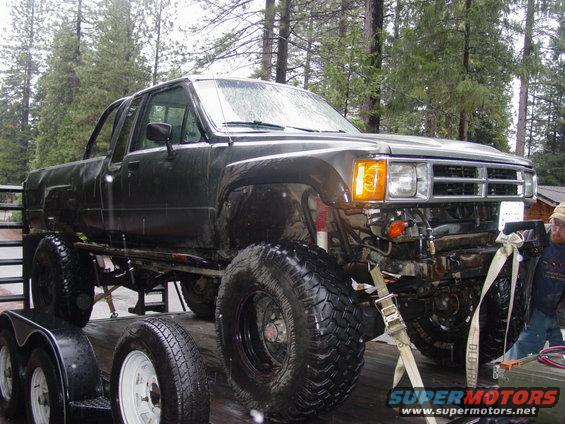 Dark green pickup truck with oversized tires on a trailer, front 3/4 view