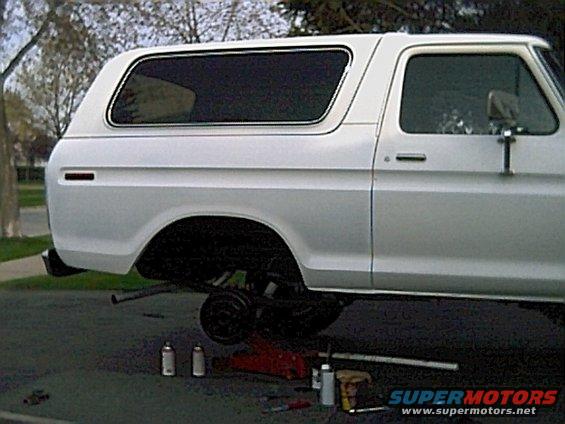 Side view of a white 1978 Ford Bronco with its rear wheel off and a jack underneath