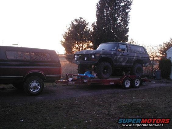 Dark Ford Bronco with camouflage pattern on a trailer, being towed by a red pickup truck