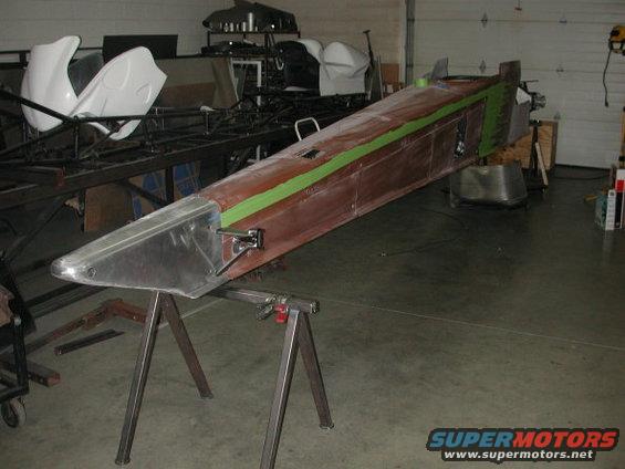 Dragster chassis with aluminum nose cone and green tape on brown bodywork, supported by metal sawhorses in a garage