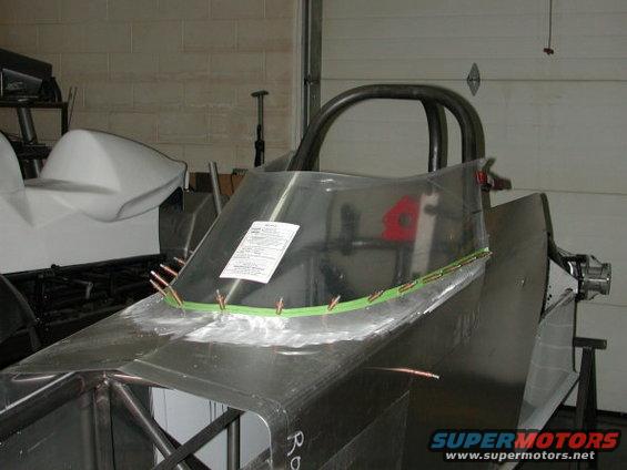 Unfinished dragster chassis with a clear windshield and roll cage, showing metal fabrication and a document attached to the windshield