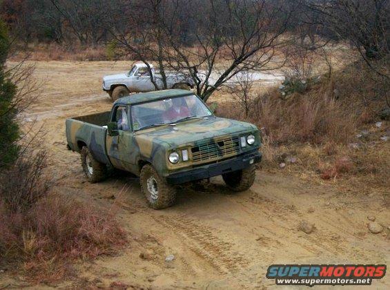Camouflage pickup truck driving on a dirt road with another white pickup truck behind it