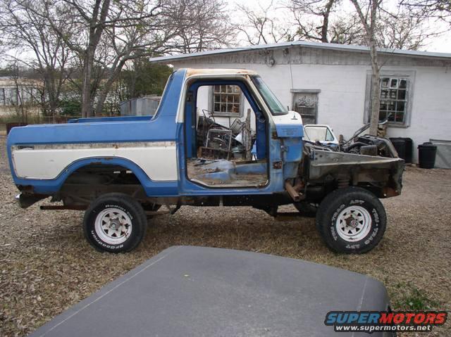 Blue and white Ford Bronco missing doors and hood, sitting in a yard