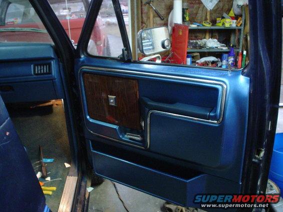 Blue interior door panel with wood grain accent and chrome trim. Another vehicle is visible through the window
