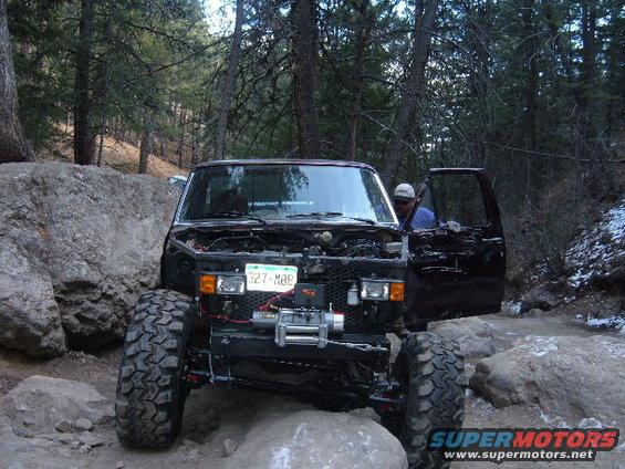 Black 1993 Ford Bronco off-roading, hood open, winch mounted, with large tires navigating rocks