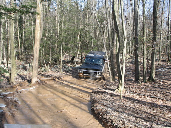 Black SUV with a prominent grille driving through a muddy trail in a forest