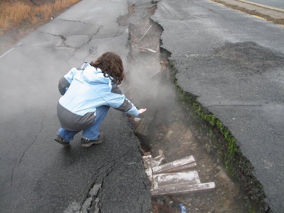 Person crouches by a large road crack with steam rising
