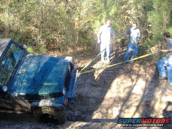 Dark green Jeep Wrangler stuck in mud, being pulled by a yellow strap by three people in a wooded area
