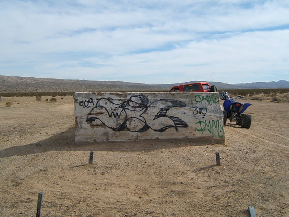 Graffiti on concrete barrier in desert landscape. Blue quad bike and red SUV visible in background