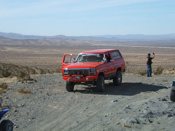 Red 1988 Ford Bronco with driver's door open, parked on a dirt road in a desert landscape. A person stands nearby, waving. Another vehicle is partially visible on the right