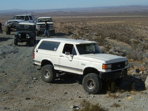 White Ford Bronco on a dirt road with other trucks behind it