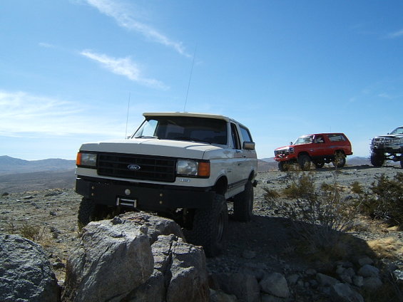 White Ford Bronco on rocks, red Bronco and white SUV in background, desert landscape