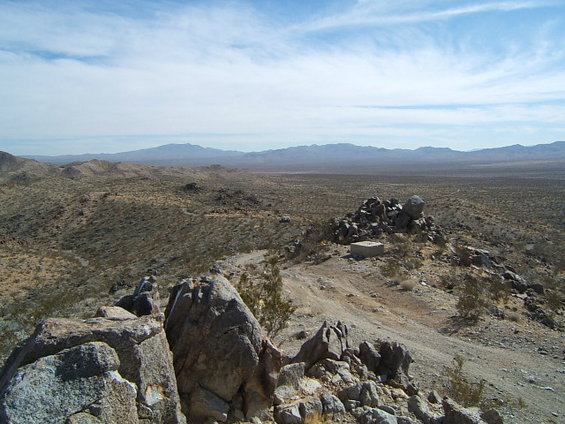 Desert landscape with rocky foreground, sparse vegetation, a small building on a hill, and distant mountains under a cloudy sky