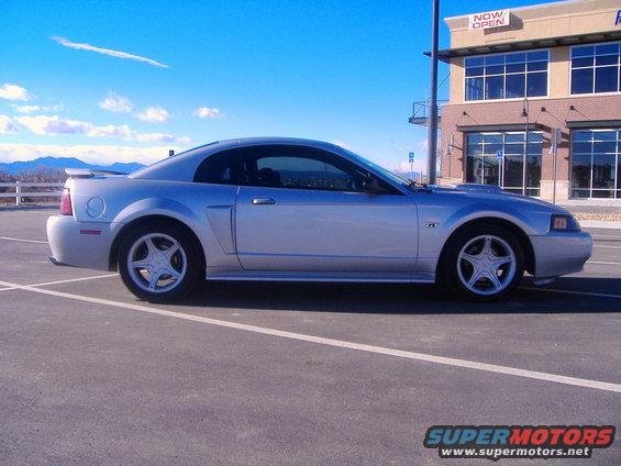 Silver 2003 Ford Mustang parked in a lot. Building in background has 'NOW OPEN' sign