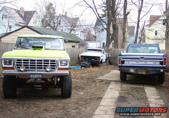 Three pickup trucks parked in a yard. The front truck is lime green with a hood scoop. A white truck is in the middle, and a blue truck is on the right