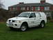 2006 Mitsubishi L200 - A white SUV with BT branding is parked on a grassy lawn in front of a brick house.
