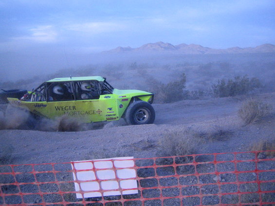 Lime green off-road buggy with roll cage and two occupants, kicking up dust on a dirt track