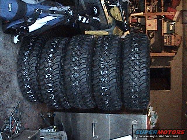 Five large off-road tires stacked up. Some have white lettering on the sidewalls