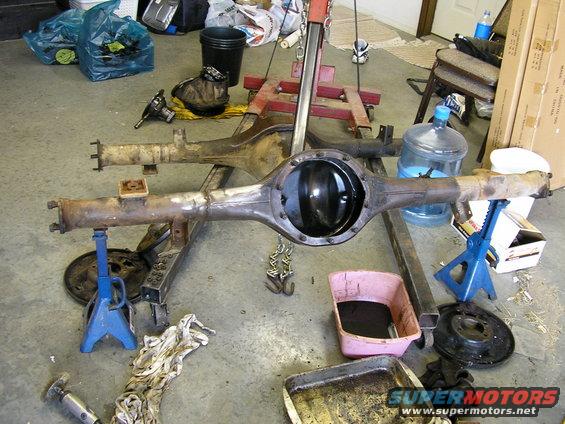 Two vehicle rear axles are on jack stands in a garage, one with the differential cover removed