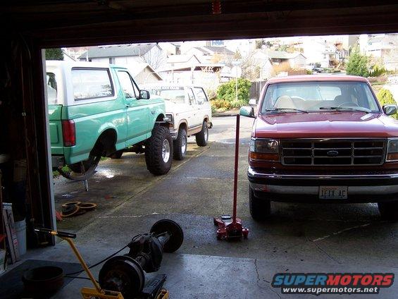 Red Ford F-150 pickup truck in front of a green Ford Bronco and a white Ford Bronco, all in a garage