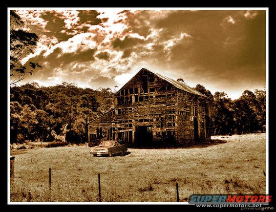 Sedan with a light-colored body and dark roof parked in a grassy field near a wooden barn