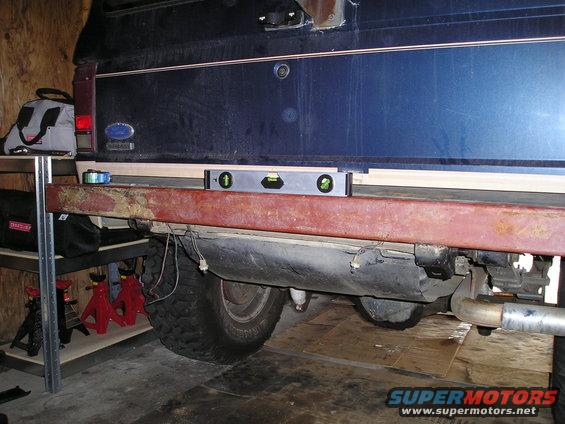 Rear of a dark blue SUV with a rusty bumper. A level sits on top of the bumper, and a fuel tank is visible underneath