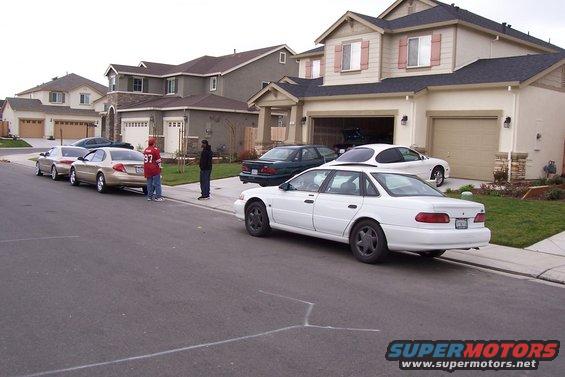 Several cars parked on street in front of houses. White sedan in foreground, gold sedan and red car behind it