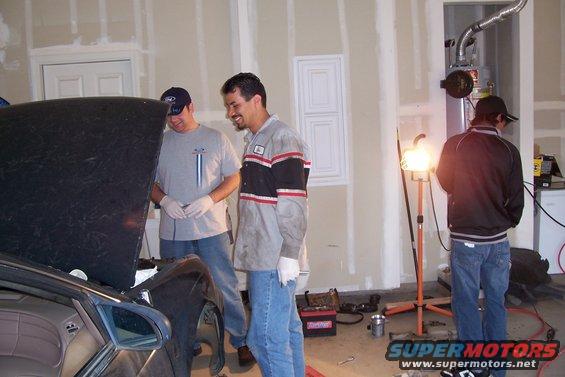Three men gathered around an open car hood in a garage, one looking at the engine