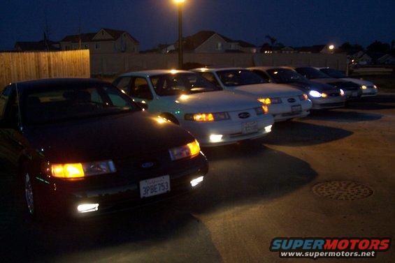 Several sedans parked in a line at night, headlights on. One car in foreground has license plate 3PBE753
