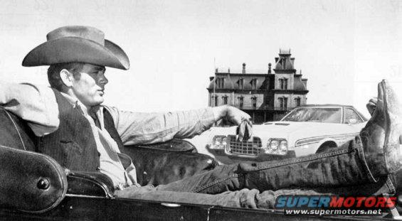 Guy in cowboy hat chilling in a car, with another car and a spooky-looking building behind him