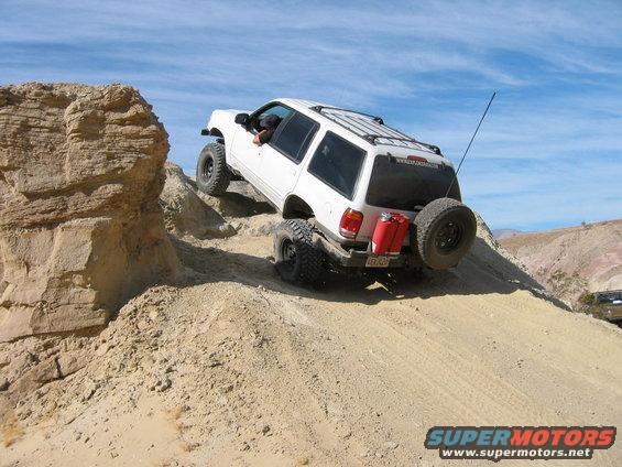 White SUV with off-road tires and spare tire on rear, climbing a sandy incline