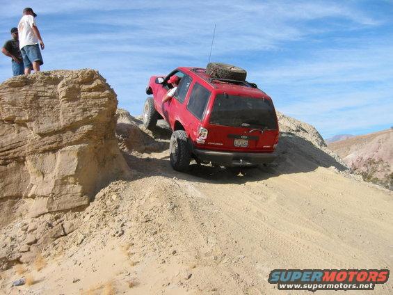 Red Ford Explorer climbing a steep sandy incline with two people watching from a rock ledge