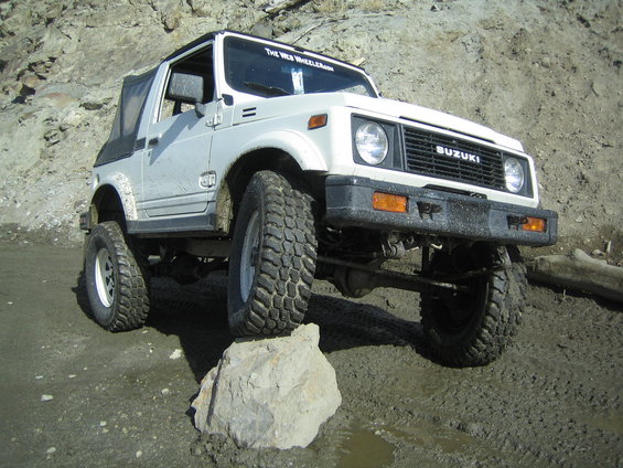 White Suzuki Samurai on a rocky, muddy trail, front tire on a rock