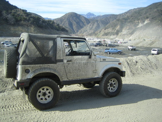 1987 Suzuki Samurai covered in dust, parked in a dry, mountainous area with other vehicles in the background