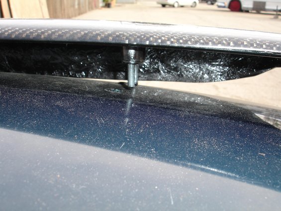 Close-up of a bolt passing through a carbon fiber spoiler and into a dark blue car body