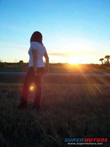 Woman in white t-shirt and jeans stands in grassy field facing away from camera towards a bright sunset over a road