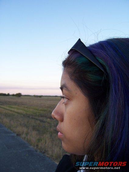 Person with colorful hair looks out at a grassy field under a pale sky. "SUPERMOTORS.NET" text is visible