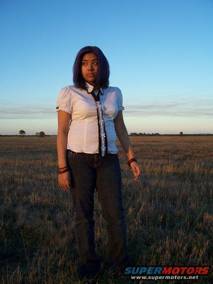 Woman with blue hair and white shirt stands in a dry field under a blue sky