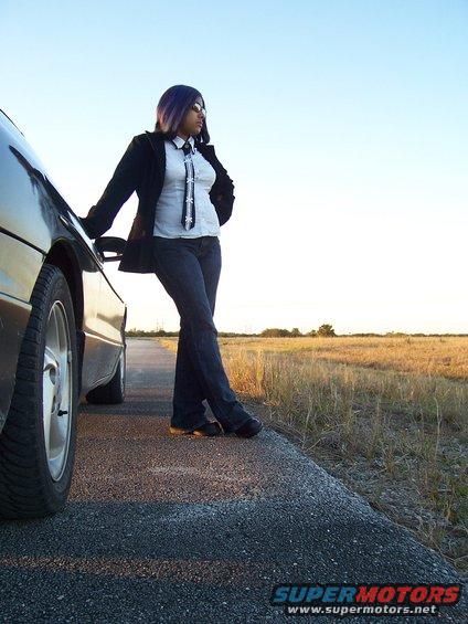 Person in a suit jacket and tie leans on the side of a car on a rural road at sunset