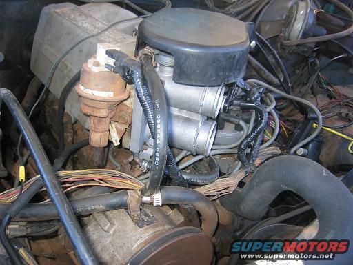 Engine bay detail showing carburetor, fuel lines, and wiring harness. Part number visible on hose