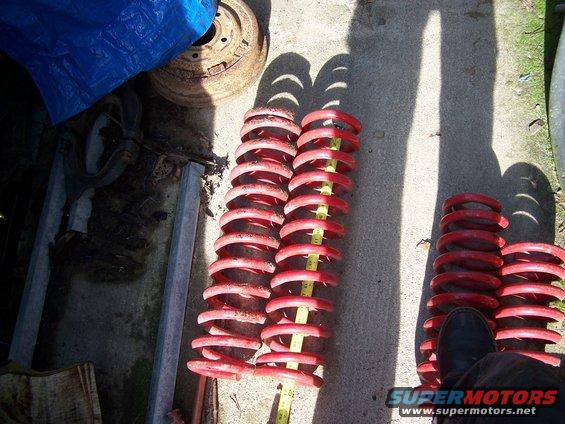 Several red coil springs laid out on concrete, one with a yellow measuring tape. A rusty wheel is visible under a blue tarp