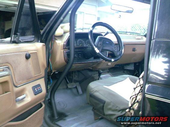 Interior of a tan and black 1987 Ford Bronco with a roll cage, aftermarket seat covers, and a black steering wheel
