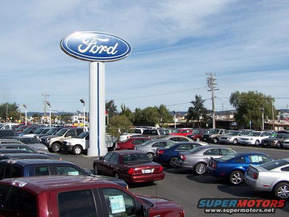 Ford dealership lot with a large Ford sign and many cars parked under a blue sky