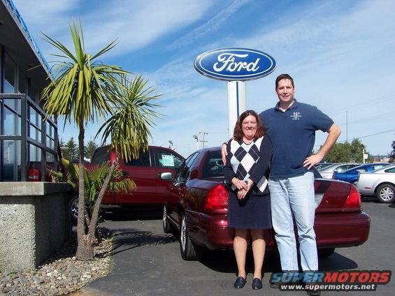 Maroon 2005 Ford Crown Victoria, rear 3/4 view, parked at a dealership with a couple standing next to it