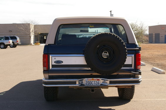 Rear view of a blue and tan Ford Bronco with a spare tire mounted on the back. Another SUV is parked to the left