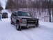 2000 Chevrolet Tahoe - A silver SUV with a bull bar is stuck in the snow, with another vehicle visible behind it on a wooded road.