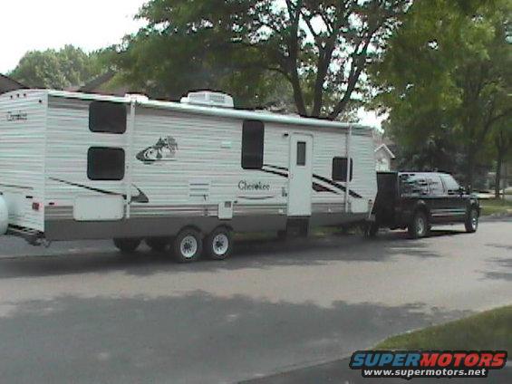 Black pickup truck towing a white Cherokee travel trailer down a residential street