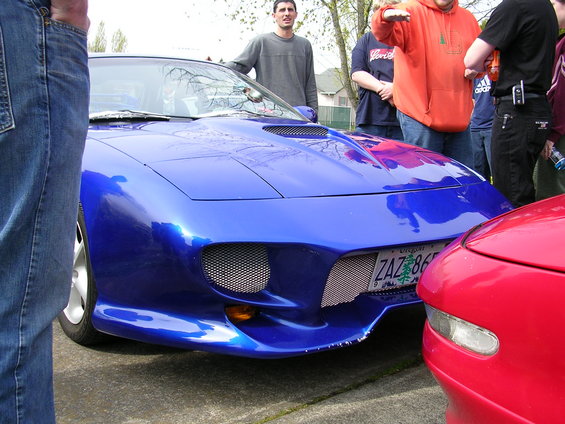 Blue sports car with custom grille and hood scoops. Red car is partially visible on the right. People stand around the cars